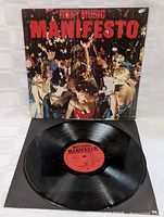 Photo shows the Roxy Music Manifesto vinyl LP in its cover, with the vinyl record displayed in front. The cover art depicts a crowd of people at a party or club scene wearing various costumes.