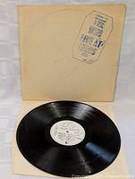 Photo showing the vinyl record outside its white cover with printed design stamp. Vinyl surface appears clean and shiny with visible grooves and label with handwritten track list.