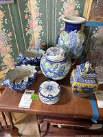 Five Asian inspired porcelain decorative pieces with blue and white patterns and one yellow and blue square container on wooden table in front of floral wallpaper.