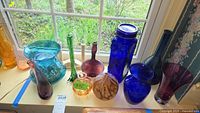 Photo showing multiple assorted colored hand blown glass vases and jar on a window sill, including cobalt blue, green, purple and amber pieces.