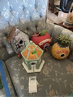 Four decorative birdhouses arranged on a floral-patterned armchair; visible apple, pineapple, signed wooden birdhouse and ceramic gazebo-shaped birdhouse