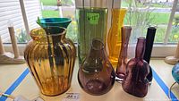 Collection of seven colored glass vases of varying shapes and colors placed on a windowsill with outdoor background.
