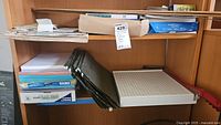 Shelf with assorted office supplies including stacked copy paper reams, black hanging file holders, a 12x12 paper cutter with red handle and grid surface, and wooden yardsticks on the top shelf.