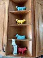 Four cow-shaped creamers or butter dishes in blue, yellow, light blue, and red on wooden shelves.