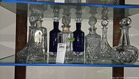 Eight assorted glass decanters displayed on glass shelves showing variety of styles, shapes, and sizes with different stoppers.
