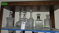 Full group photo showing six different glass decanters arranged on glass shelf under wood frame, various sizes and shapes visible.