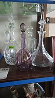 Four glass decanters of varying shapes and sizes with stoppers on a wooden shelf.