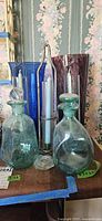 Two green glass decanters in front, tall blue and purple glass vases behind, two metal candlestick holders with light blue taper candles in between. Items show some dust and need cleaning.