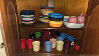 Image showing stacks of colorful Fiesta Ware dinner plates and small bowls on top shelves and a row of various colorful mugs and cups on bottom shelf inside a wooden cabinet.