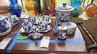 Complete china set on wooden table includes cups, saucers, biscuit jar, trinket box, bird figurine; multi-colored glass vases in background
