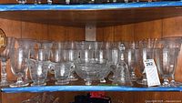 Shelf display of multiple Fostoria etched glass footed glasses, a creamer, sugar bowl, a footed bowl, and a glass bell.