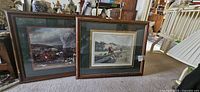 Two wood framed fox hunt prints shown propped against furniture indoors, revealing overall condition and size.