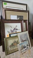 Photo showing a stack of 8 framed artworks with visible country scenes, ducks, houses, and winter landscapes, arranged leaning against a wall.