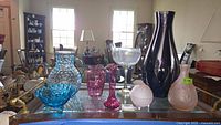View of the lot on glass table showing a blue hobnail pitcher and dish, two pink frosted glass pieces, two purple vases, and a large printed wine glass.
