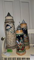 Three German beer steins of varying sizes on a white surface. The largest is beige and blue with a raised relief of a group of men. Two smaller navy blue steins portray painted figures in traditional clothing with pewter lids.