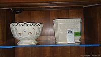 Two Lenox ceramic items on a glass shelf: the Peony Basket bowl left with floral designs and open basket weave, and the white Forum ice bucket on the right with decorative classical motifs.