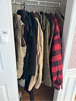 Closet view showing men's jackets and vests hanging on a single rack, including a red and black plaid jacket, tan vest, and other neutrally colored outerwear.