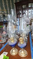 Photo showing collection of vintage oil lamps in clear and blue glass, with brass burner elements and chimneys, arranged on a wooden table.