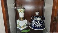Covered figural cheese or butter dish and Italian porcelain pepper mill displayed on wooden shelf. The pepper mill is square shaped with floral decorations and brass top, approx 8.5-9.5 inches tall. The covered dish is predominantly blue and white with figural hand-painted dotted and circular pattern.