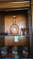 Photo of wooden cabinet shelf displaying pink glass decanter, two matching pink glasses, small amber hobnail vase, ashtray, and three hand painted koi fish glasses on lower shelf.