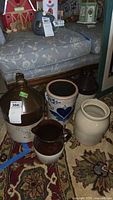 Photo showing four pottery items on a rug in front of a cushioned bench; items include two large jugs, one decorated crock with blue heart, and one plain beige crock.