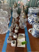 Photo showing an assortment of salt and pepper shakers of various sizes and materials including glass and ceramic with silver tops, along with other blue and white ceramic items in the background.