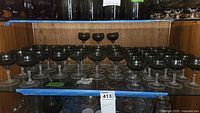 Multiple Sasaki crystal stemmed glasses with smoky grey tint arranged on wooden shelves, total of 25 glasses visible.