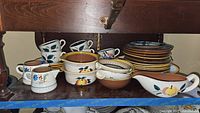Photo showing stacked Stangl pottery cups, creamers, plates, and bowls with painted fruit and floral designs. Visible signs of use and nicks.