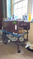 Tea cart fully assembled showing side with teaware on top and plates on second and third shelves, highlighting wheels and handle.
