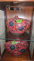 Two red Asian themed vases with black and white floral and vine designs, placed on two glass shelves inside a wooden display cabinet, showing full view of both vases with size tags visible