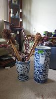 View of two blue and white porcelain items on carpet; left is floral decor planter with dried artificial flowers, right is taller umbrella stand; both items have floral blue designs on white background.