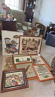 Photo showing multiple framed needlepoint artworks arranged on floor, featuring scenes of houses, animals, and other themes.