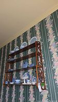 Full view of vintage wooden wall-mounted shelf with decorative carved sides, containing various blue and white porcelain plates and cups (not included in sale). Shelf hung on floral wallpapered wall.