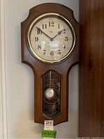 Front view of Elgin battery operated pendulum wood wall clock showing clock face and brass bezel with visible pendulum and decorative glass panel below.