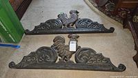 Two wooden carved rooster wall mount pieces shown side by side on carpeted floor with ornate scrollwork and rooster central figures, dark wood finish.