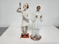 Photo showing two Goebel ceramic figurines: a man holding a bottle standing on a square base, and a figure in a white robe with mosaic floral base.