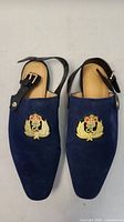 Top view of both blue suede slingback shoes with gold embroidered crest and black leather ankle straps.