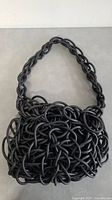 Black purse constructed from intertwined black wire tubes forming a textured mesh surface with a thick linked strap.