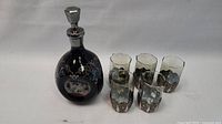 Photo showing full lot of black glass decanter with silver-tone metal overlay and four matching glasses with metal holders arranged around it.
