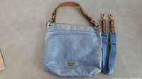 Light blue leather FOSSIL purse laying flat with leather handle beside detachable blue fabric shoulder strap with leather trim and gold-tone hardware hooks.