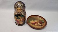 Photo showing a single Matryoshka doll with detailed traditional artwork alongside the wooden plate featuring painted peaches.
