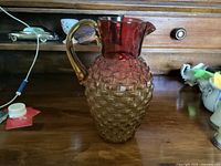 Amberina glass pitcher on wood surface showing its textured diamond pattern and red to amber gradient coloring with handle.