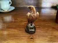 Front view of the brown ceramic eagle figurine on wooden surface with some ceramic cups in the background, showing upright eagle perched on base with detailed feathers.