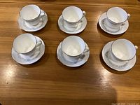 Top down view of six white porcelain cups with handles, each paired with a matching saucer on a wood surface.