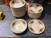 Stack of 21 cereal bowls and 16 bread plates with pink and red rose floral design, sitting on a blue countertop.