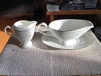 Gravy boat with matching underplate and creamer set, shown on textured surface with sunlight highlighting fine details.
