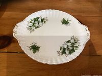 Top view of the white porcelain cake plate with trillium floral decorations and gold trimmed scalloped edges and handles.