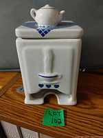Front view of white ceramic tea bag holder shaped like a stove, showing blue heart accents and a small teapot on the lid.
