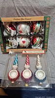 Photo showing two unopened boxes containing Christmas glass ornaments. One box contains six round ornaments decorated with red, white, and green holiday motifs. The other box has three pointed finial-style ornaments in red, blue, and white with swirls and shiny reflective centers.