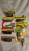 Photo of assorted packaged fishing lures dominated by CULPRIT brand soft plastics in various colors such as brown and yellow, and a YUM soft plastic lure.